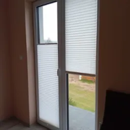 Białe, plisowane osłony okienne zamontowane na dwuskrzydłowych drzwiach balkonowych z widokiem na ogród.