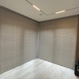 GREEN HOUSE Plejada-Stary Nowy Dom - Jasne, aluminiowe żaluzje poziome zamontowane w rogu pokoju z drewnianą podłogą i oświetleniem LED na suficie.