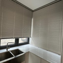 GREEN HOUSE Plejada-Stary Nowy Dom - Szare poziome lamele w oknach kuchni, widok na zewnątrz częściowo zasłonięty, nowoczesna aranżacja z metalowym zlewem i jasnym blatem.