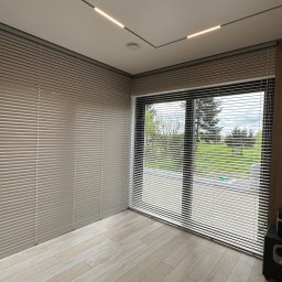 GREEN HOUSE Plejada-Stary Nowy Dom - Jasne, aluminiowe żaluzje poziome zamontowane w rogu pokoju, częściowo zasłonięte, z widokiem na zewnątrz przez okno. Nowoczesne oświetlenie sufitowe.