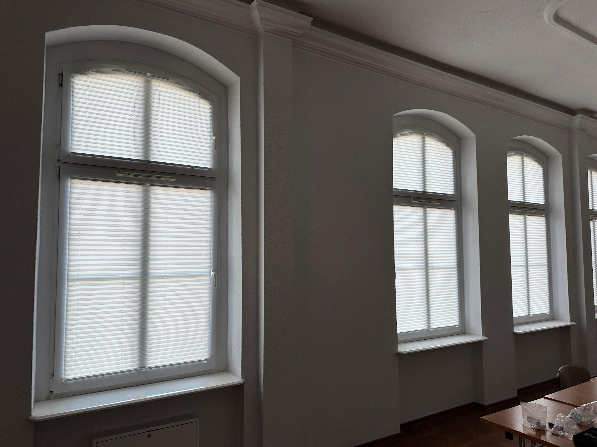 Trzy okna z białymi, plisowanymi osłonami okiennymi w klasycznym wnętrzu z listwami przysufitowymi.