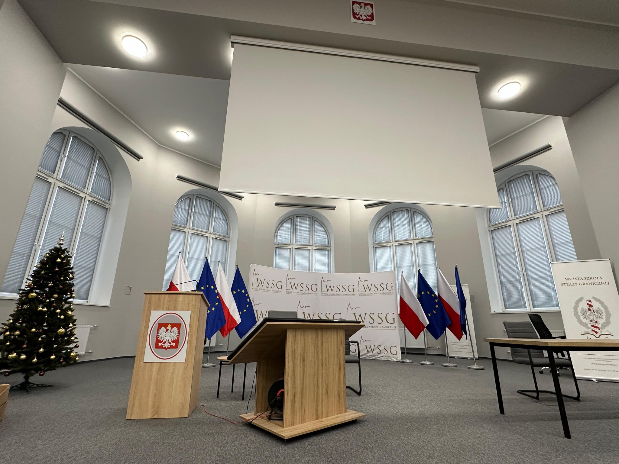 Sala konferencyjna z podniesionym ekranem projekcyjnym, mównicą z godłem Polski, flagami Polski i Unii Europejskiej oraz choinką w rogu
