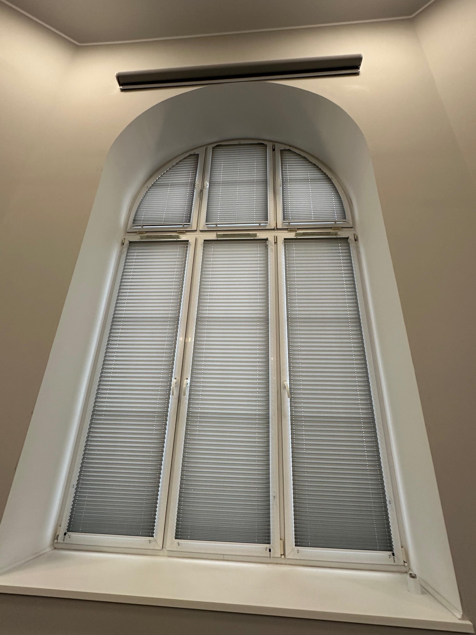 Wysokie okno łukowe z białymi ramami, wyposażone w szare plisy, widok z dołu.