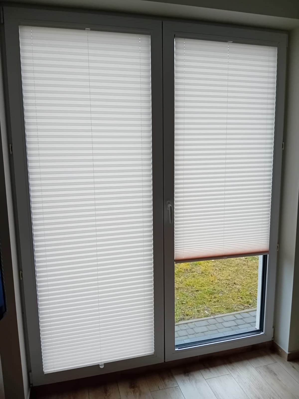 Białe plisy okienne zamontowane w oknach w Choroszczy, z widokiem na trawnik i chodnik na zewnątrz.