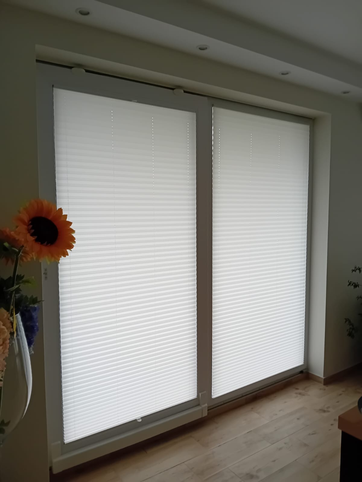 Białe plisy okienne zamontowane na dwóch oknach, widok z wnętrza, z boku dekoracyjny bukiet sztucznych kwiatów.