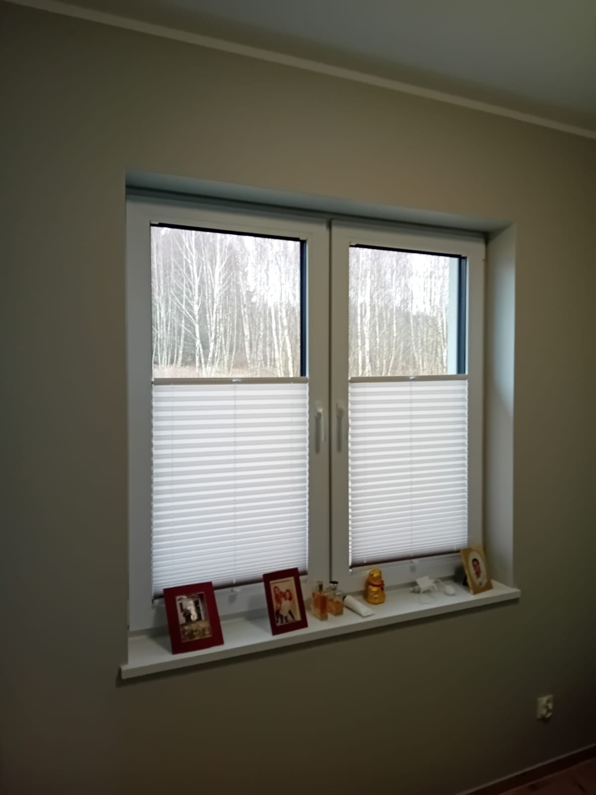 Dwuokienne okno z białymi ramami, wyposażone w częściowo zasłonięte, plisowane osłony przeciwsłoneczne. Na parapecie stoją ramki ze zdjęciami, flakoniki perfum i figurka Buddy.