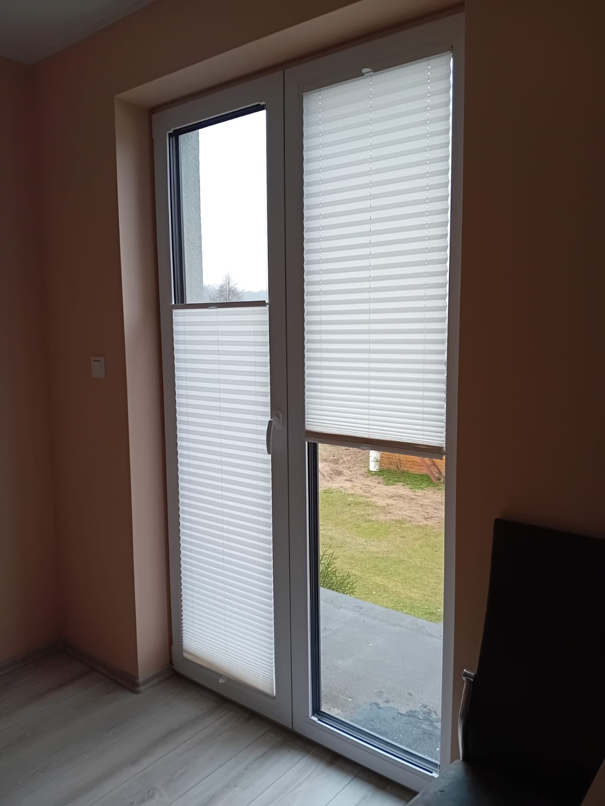 Białe, plisowane osłony okienne zamontowane na dwuskrzydłowych drzwiach balkonowych z widokiem na ogród.