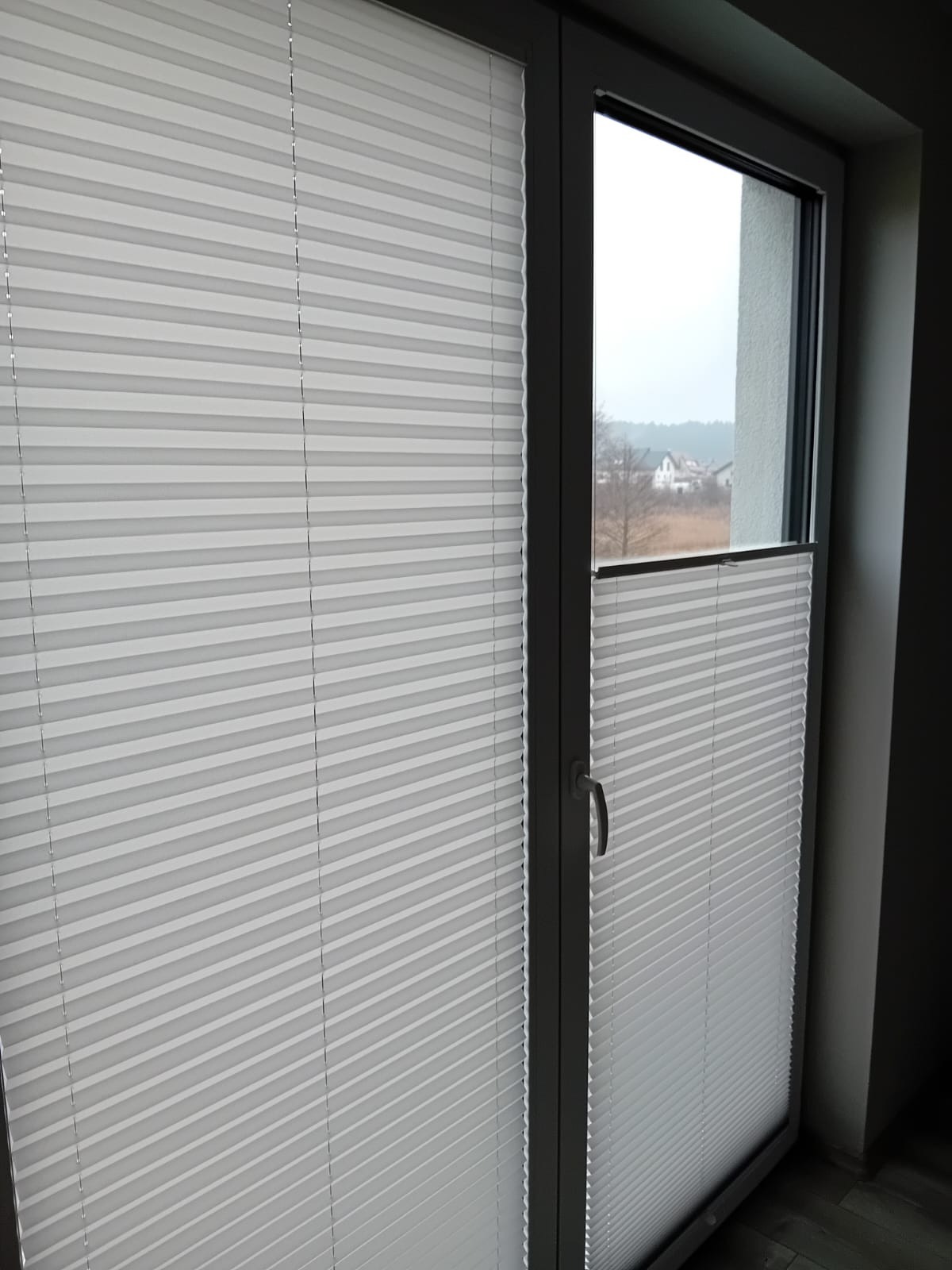 Białe, plisowane osłony okienne zamontowane w szarym oknie, częściowo zasłonięte, widok na oddalone domy i drzewa za oknem.