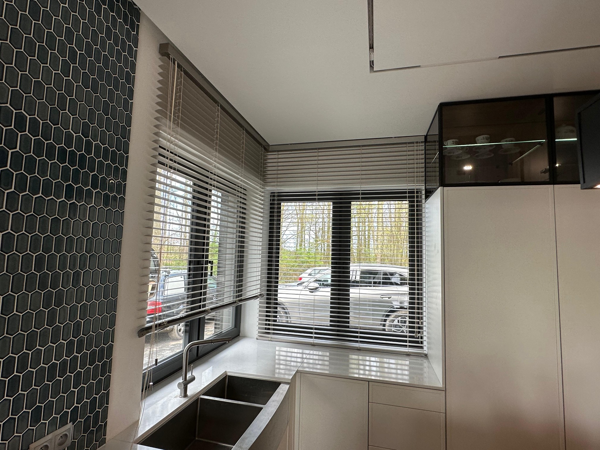 Szare żaluzje aluminiowe zamontowane w oknach kuchni z widokiem na zaparkowane samochody, widoczne heksagonalne płytki na ścianie i stalowy zlew.