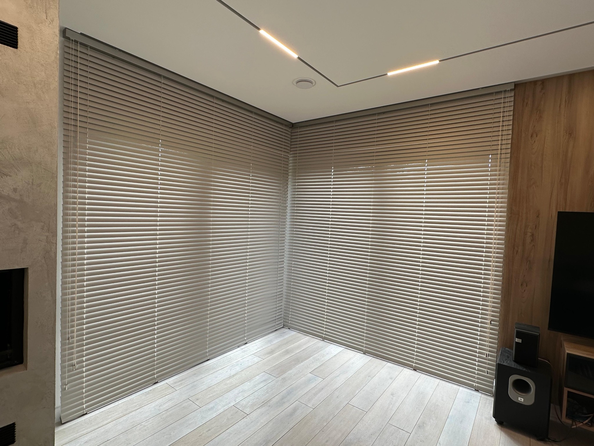 Jasne, aluminiowe żaluzje poziome zamontowane w rogu pokoju z drewnianą podłogą i oświetleniem LED na suficie.