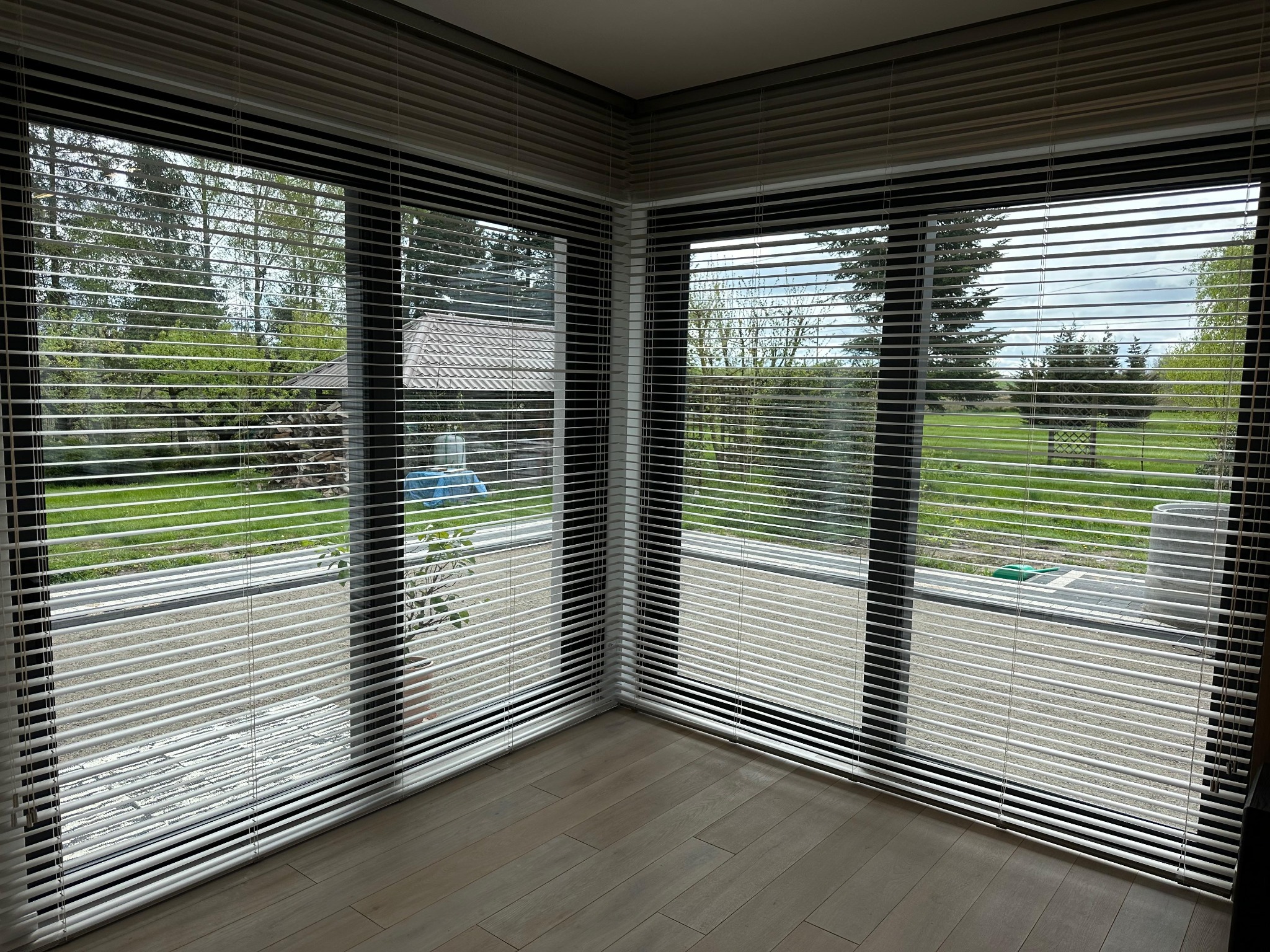 Jasne, nowoczesne wnętrze z dużymi oknami, na których zamontowano poziome żaluzje aluminiowe, częściowo zasłonięte, przez które widać zielony ogród i dom.