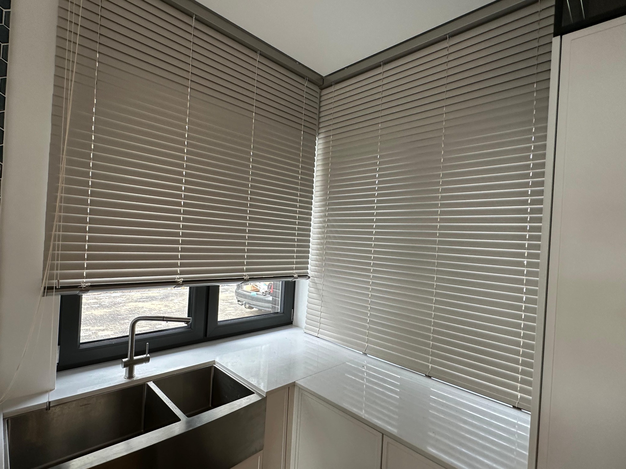 Szare poziome lamele w oknach kuchni, widok na zewnątrz częściowo zasłonięty, nowoczesna aranżacja z metalowym zlewem i jasnym blatem.