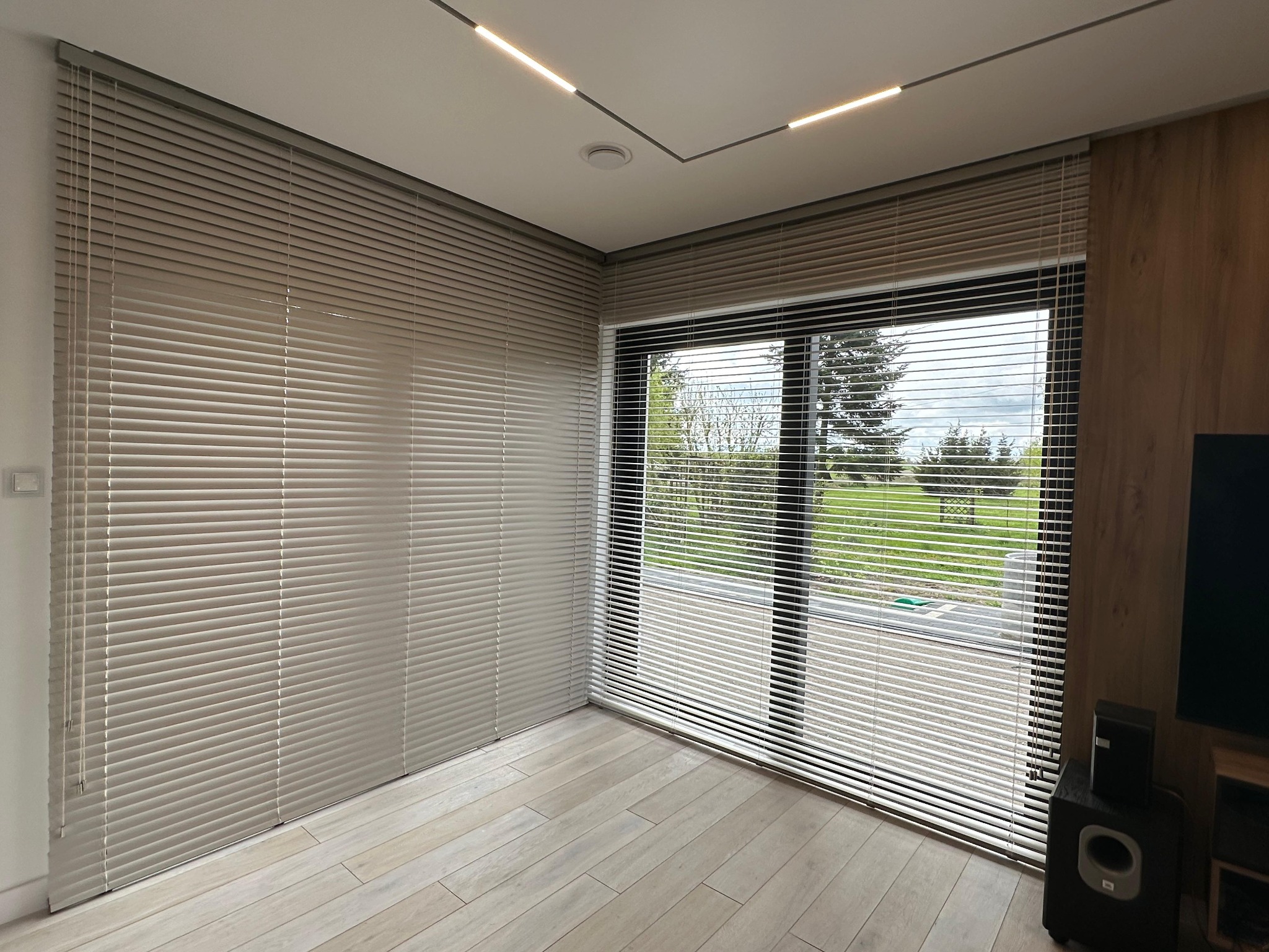 Jasne, aluminiowe żaluzje poziome zamontowane w rogu pokoju, częściowo zasłonięte, z widokiem na zewnątrz przez okno. Nowoczesne oświetlenie sufitowe.