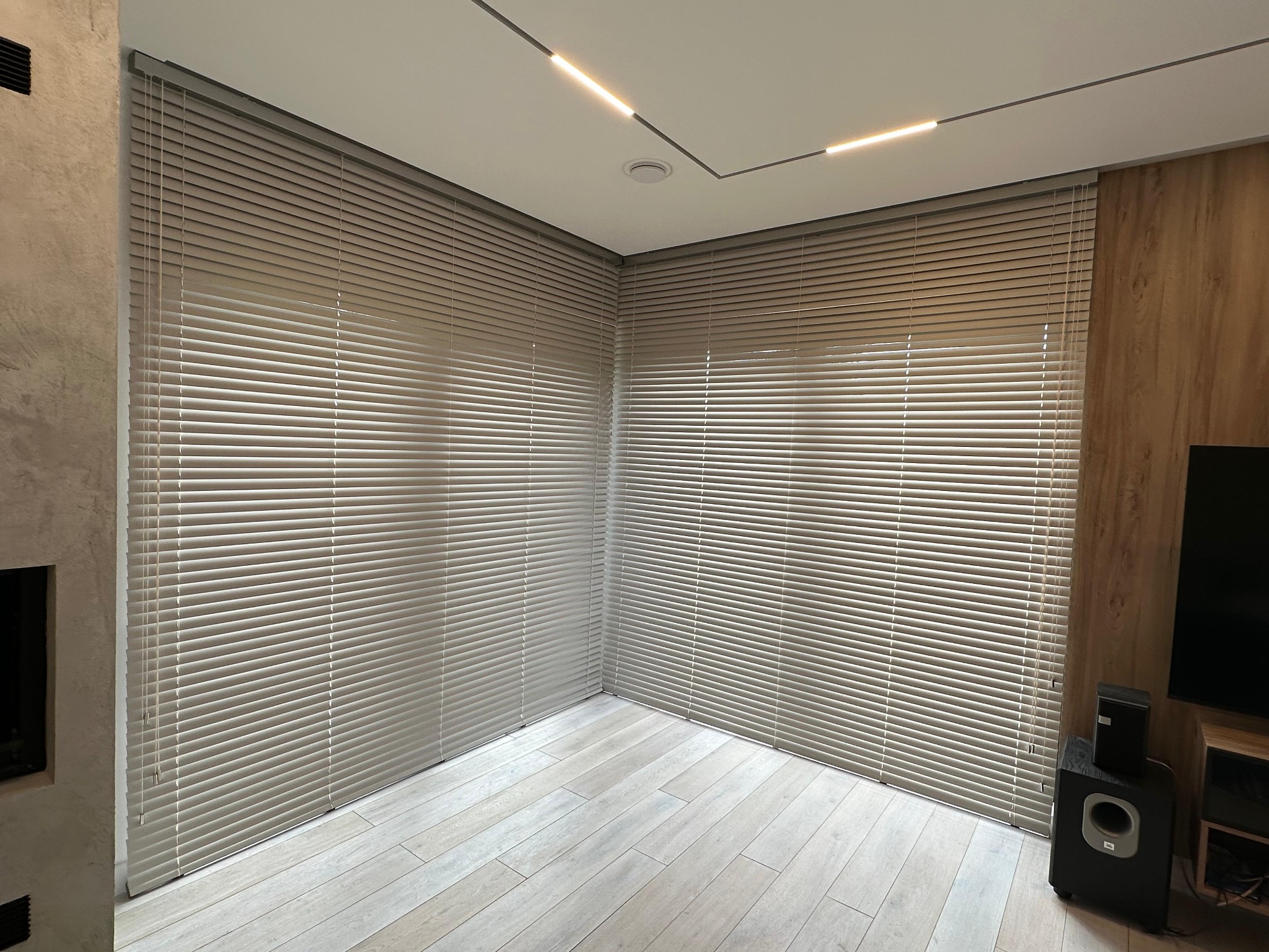 Beżowe żaluzje poziome zamontowane w rogu pokoju, widoczne listwy oświetleniowe na suficie i podłoga z paneli w odcieniach szarości.