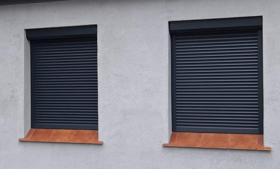 Dwa okna z całkowicie zasłoniętymi, ciemnoszarymi roletami zewnętrznymi na tle jasnoszarej, chropowatej elewacji budynku z brązowymi parapetami.