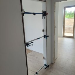 Usługi Ogólnobudowlane i Wykończeniowe Kamil Zakrzewski - Montaż ościeżnicy drzwiowej w Olsztynku, zabezpieczonej rozpórkami, widok na jasne pomieszczenie z oknem.