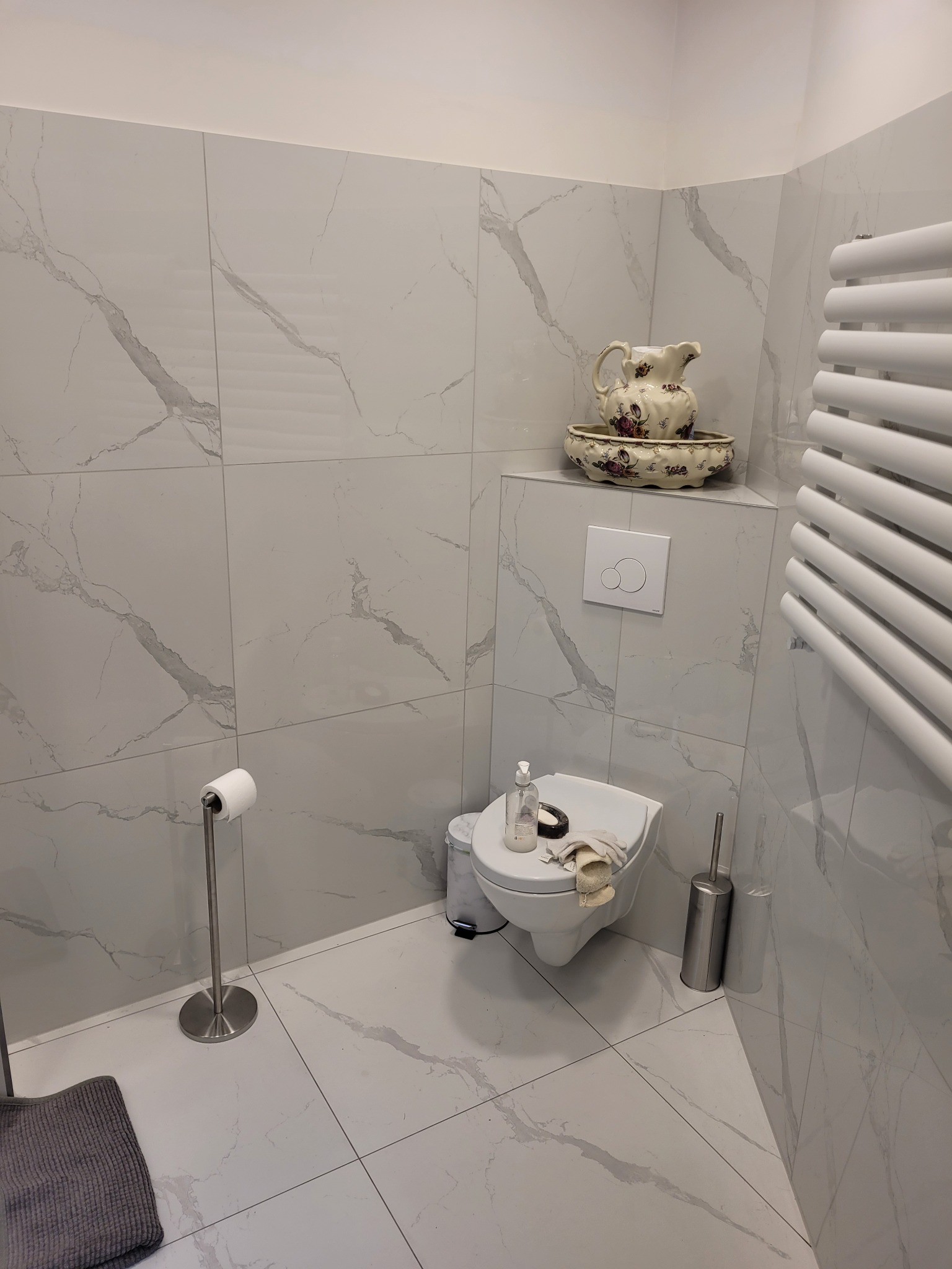 Biała łazienka z marmurowymi płytkami, wiszącą toaletą z deską, ozdobnym dzbankiem i grzejnikiem drabinkowym.