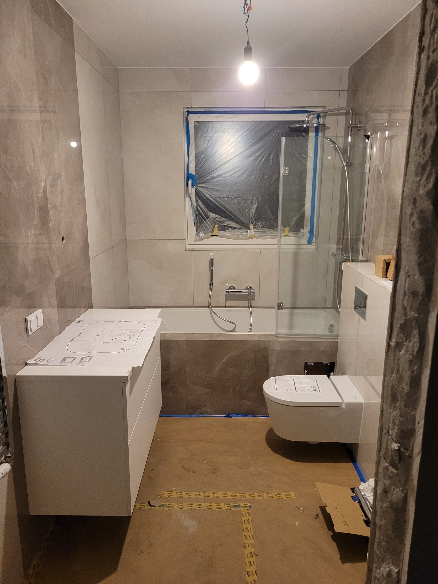 Wnętrze łazienki w trakcie remontu z częściowo położonymi płytkami, zakrytym oknem folią, podwieszaną toaletą, wanną z parawanem oraz umywalką z szafką, na której leży projekt techniczny.