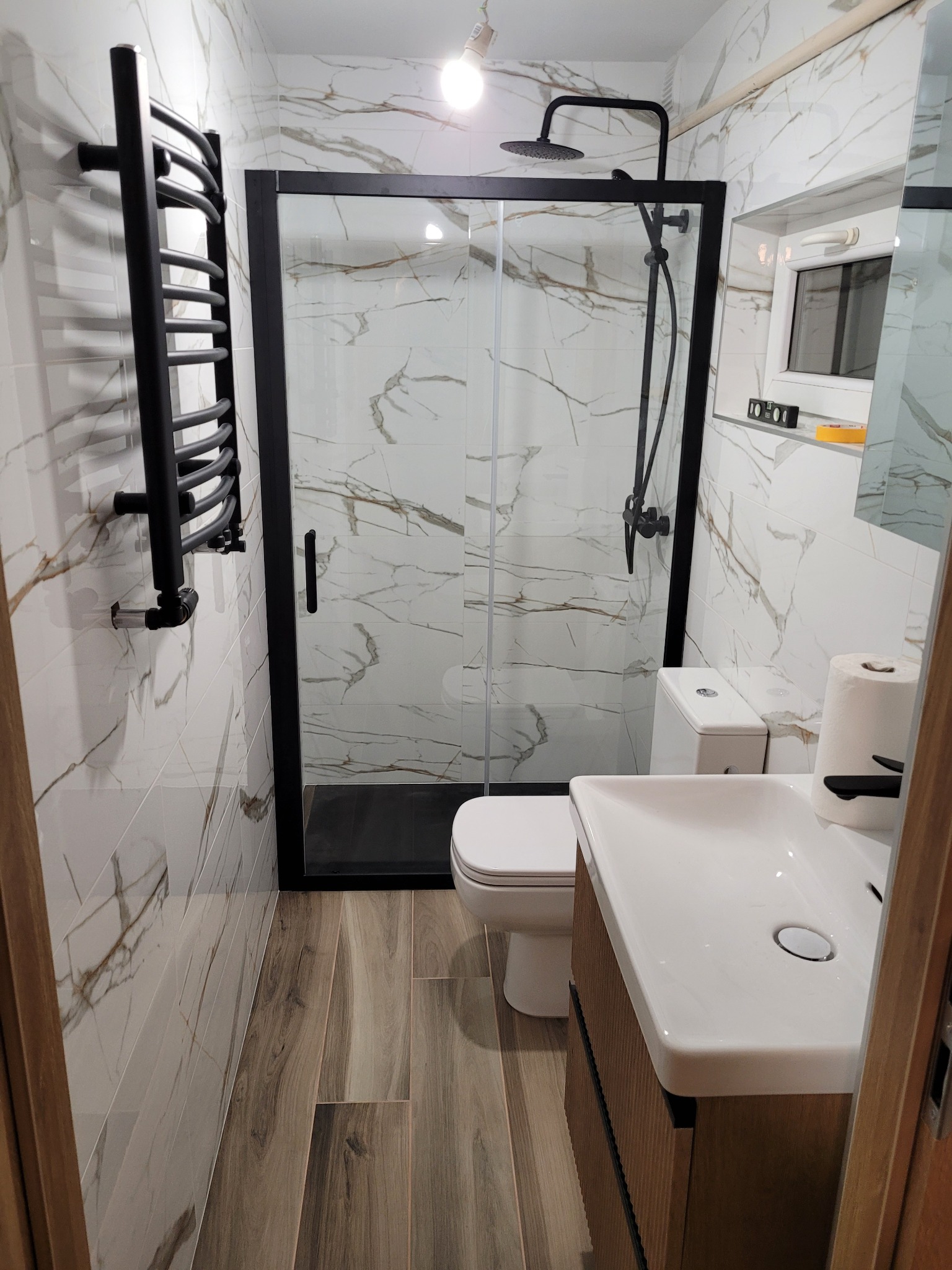 Nowoczesna łazienka z kabiną prysznicową typu walk-in z czarnymi ramami, grzejnikiem drabinkowym i umywalką z szafką w kolorze drewna; płytki imitujące marmur na ścianach i drewnopodobne na podłodze.