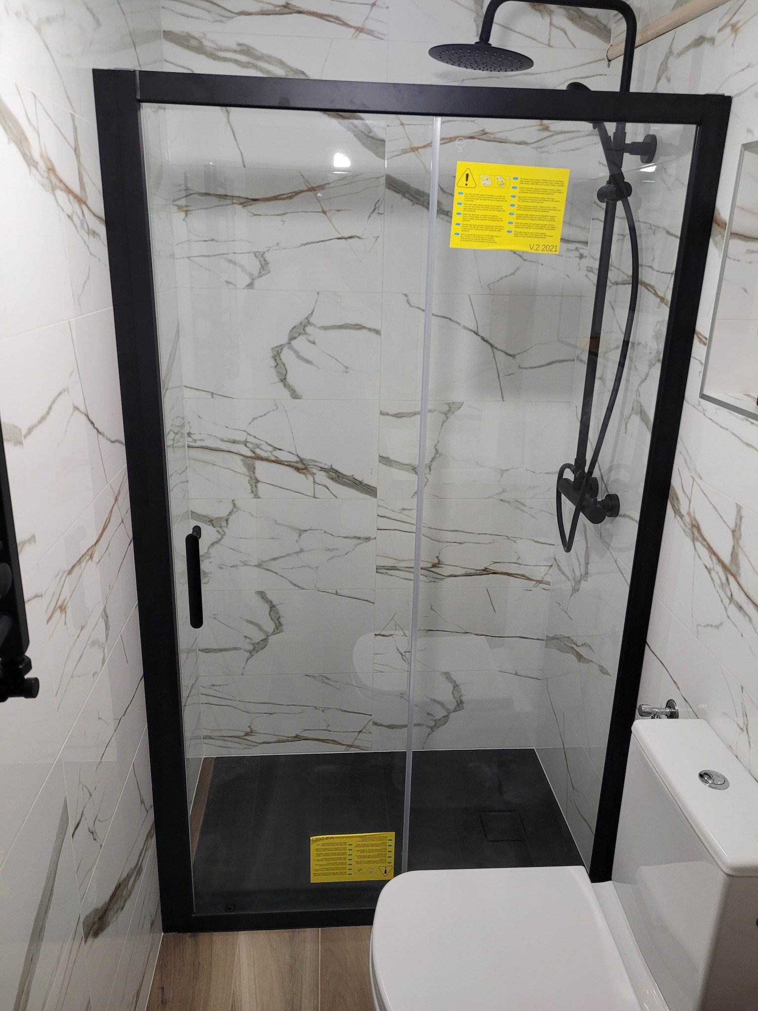 Nowoczesna kabina prysznicowa z czarną ramą i deszczownicą, wykończona płytkami imitującymi marmur, widoczna toaleta.