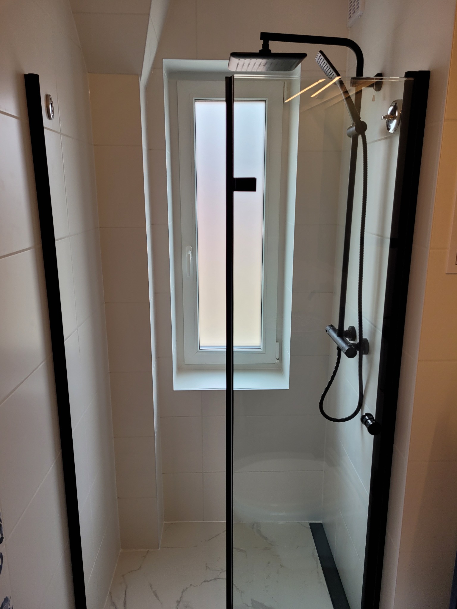 Nowoczesna kabina prysznicowa z czarnymi profilami i deszczownicą, zamontowana w łazience z oknem i białymi płytkami.