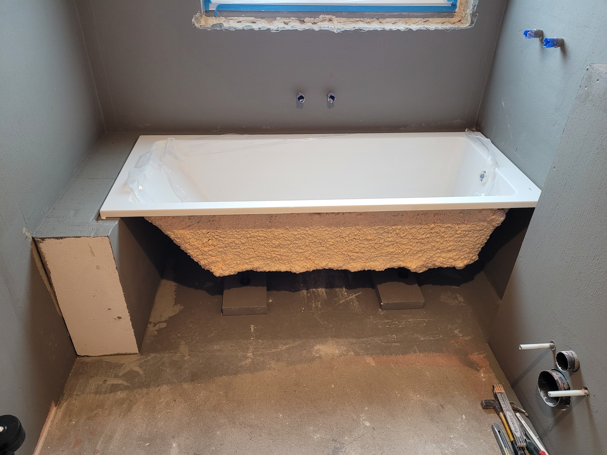 Biała wanna akrylowa, częściowo obłożona pianką montażową, ustawiona na betonowych bloczkach w surowej łazience z szarymi ścianami i widocznymi instalacjami wodnymi.