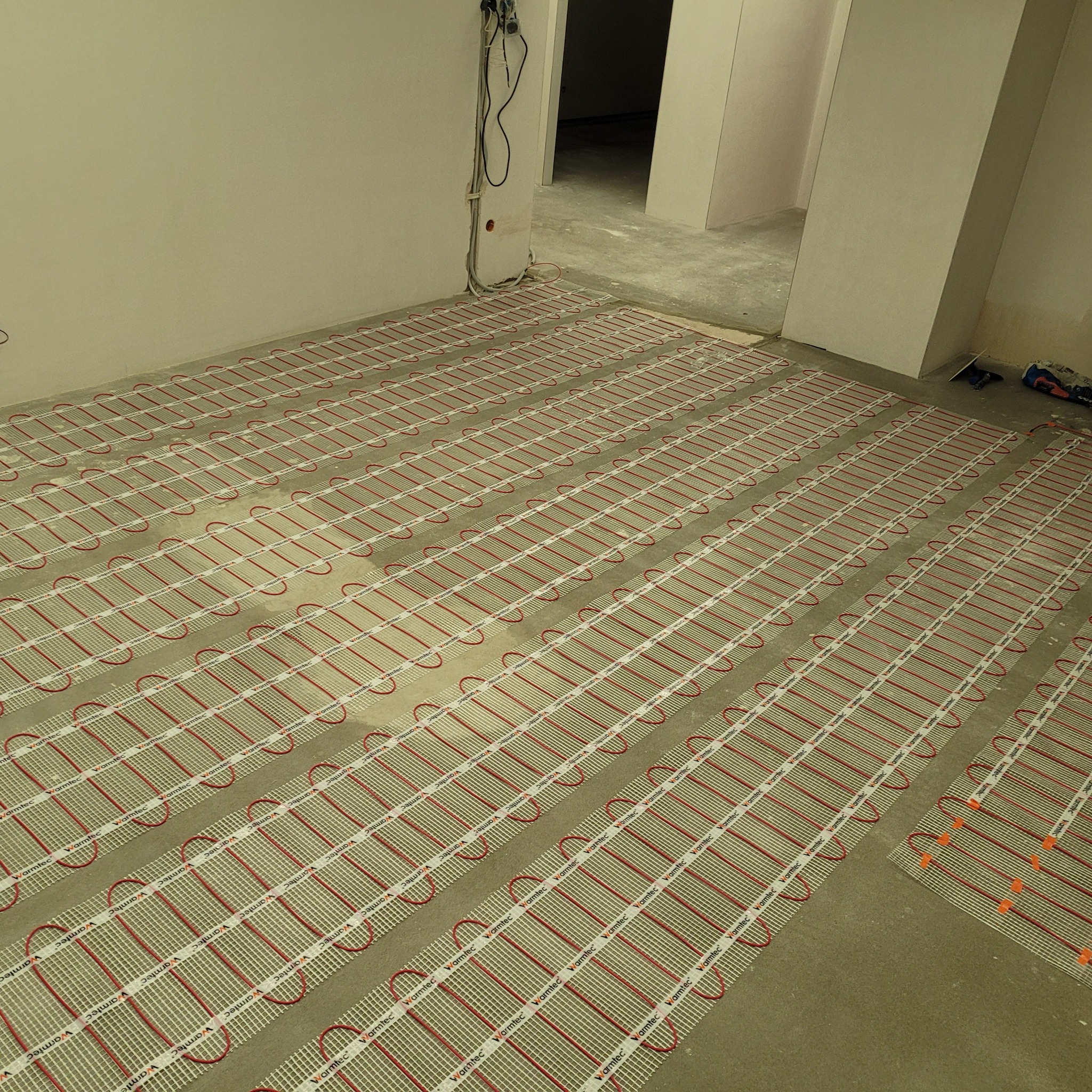 Rozłożona mata grzewcza Warmtec z czerwonymi przewodami na surowej podłodze w trakcie prac wykończeniowych, widoczne kable elektryczne wystające ze ściany.