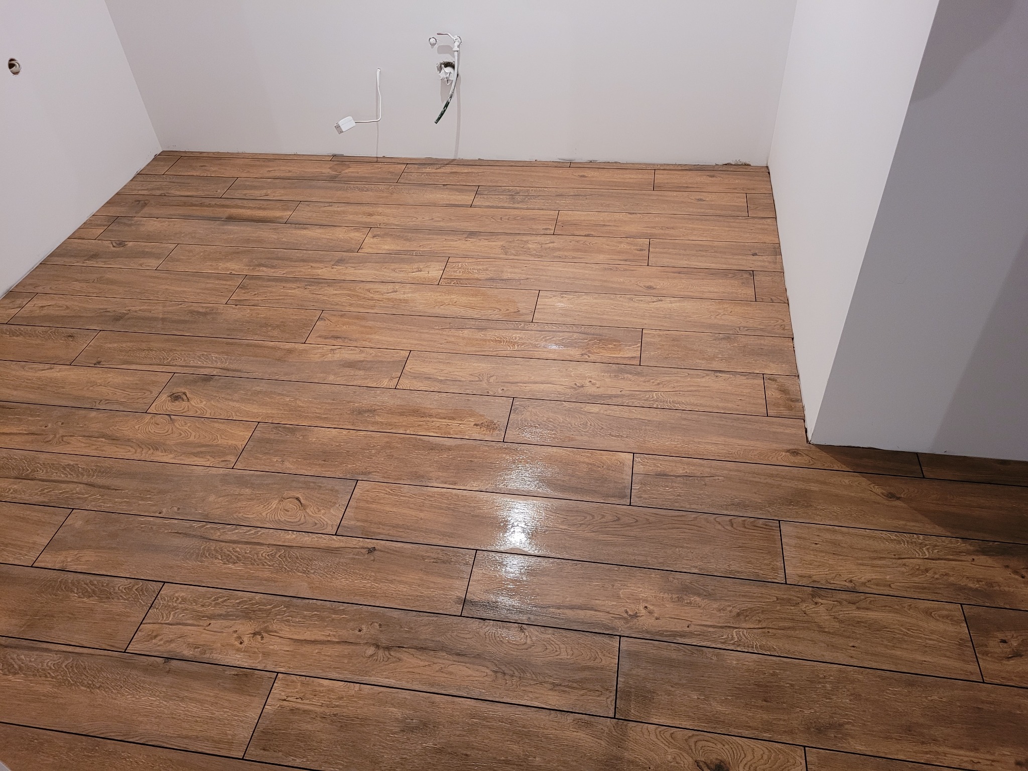 Ułożona, brązowa podłoga z płytek imitujących drewno w pustym, białym pomieszczeniu, widoczne gniazdka elektryczne na ścianie.
