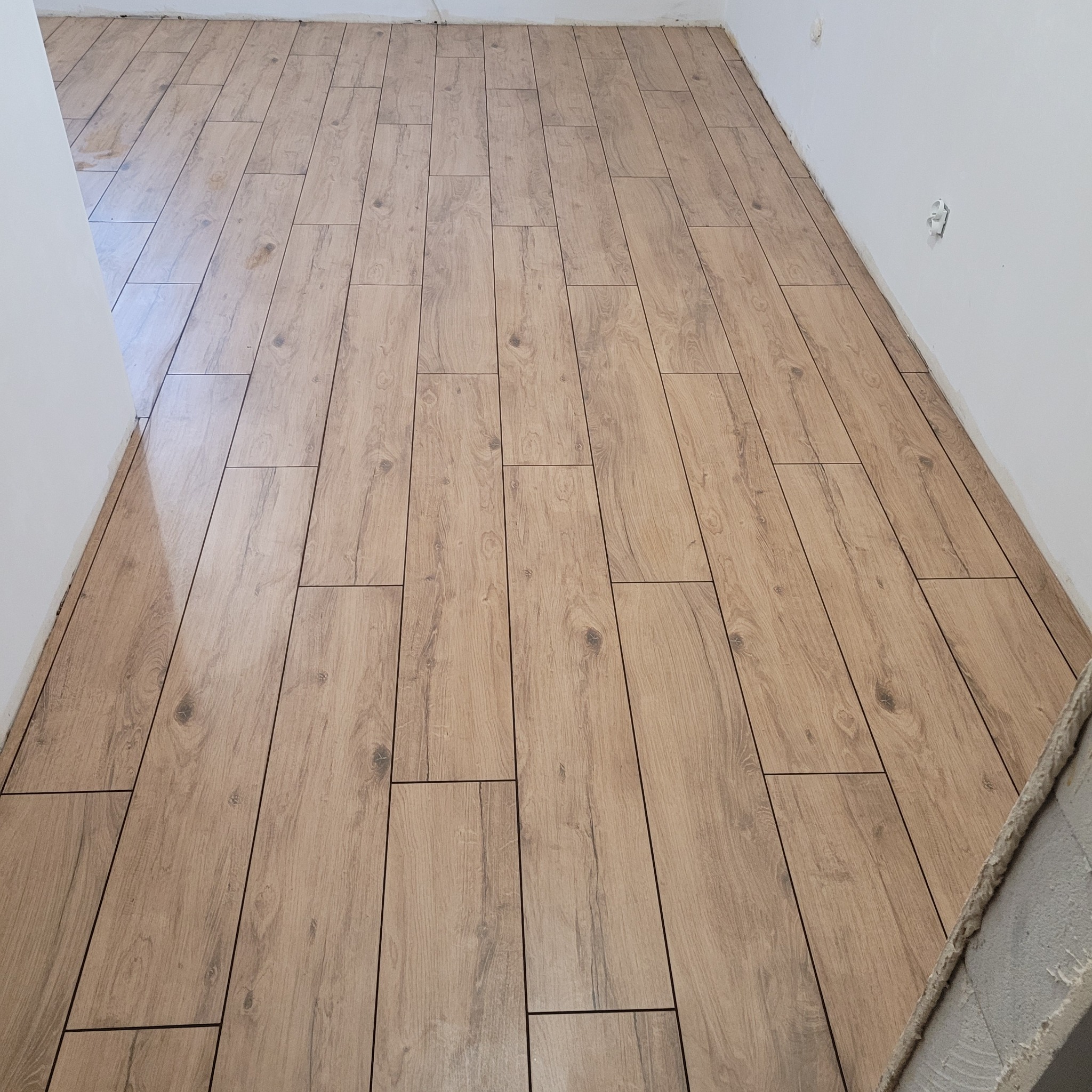 Ułożona podłoga z płytek ceramicznych imitujących drewno, widok z góry, fragment ściany z widocznymi gniazdkami elektrycznymi, świeżo położona podłoga.