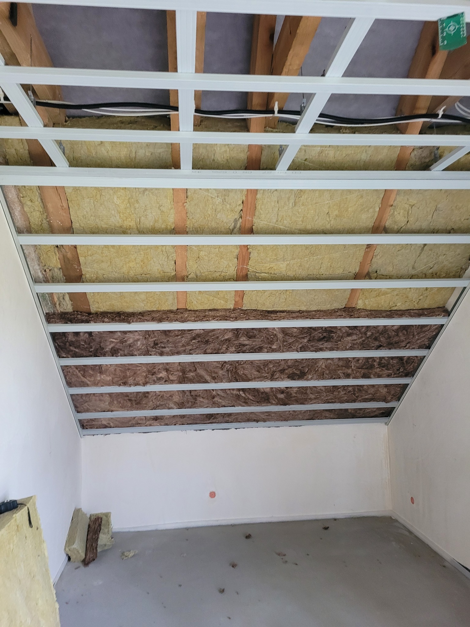 Montaż sufitu podwieszanego z izolacją z wełny mineralnej w pomieszczeniu ze ścianami pomalowanymi na biało, widoczne profile metalowe i dwa rodzaje izolacji.