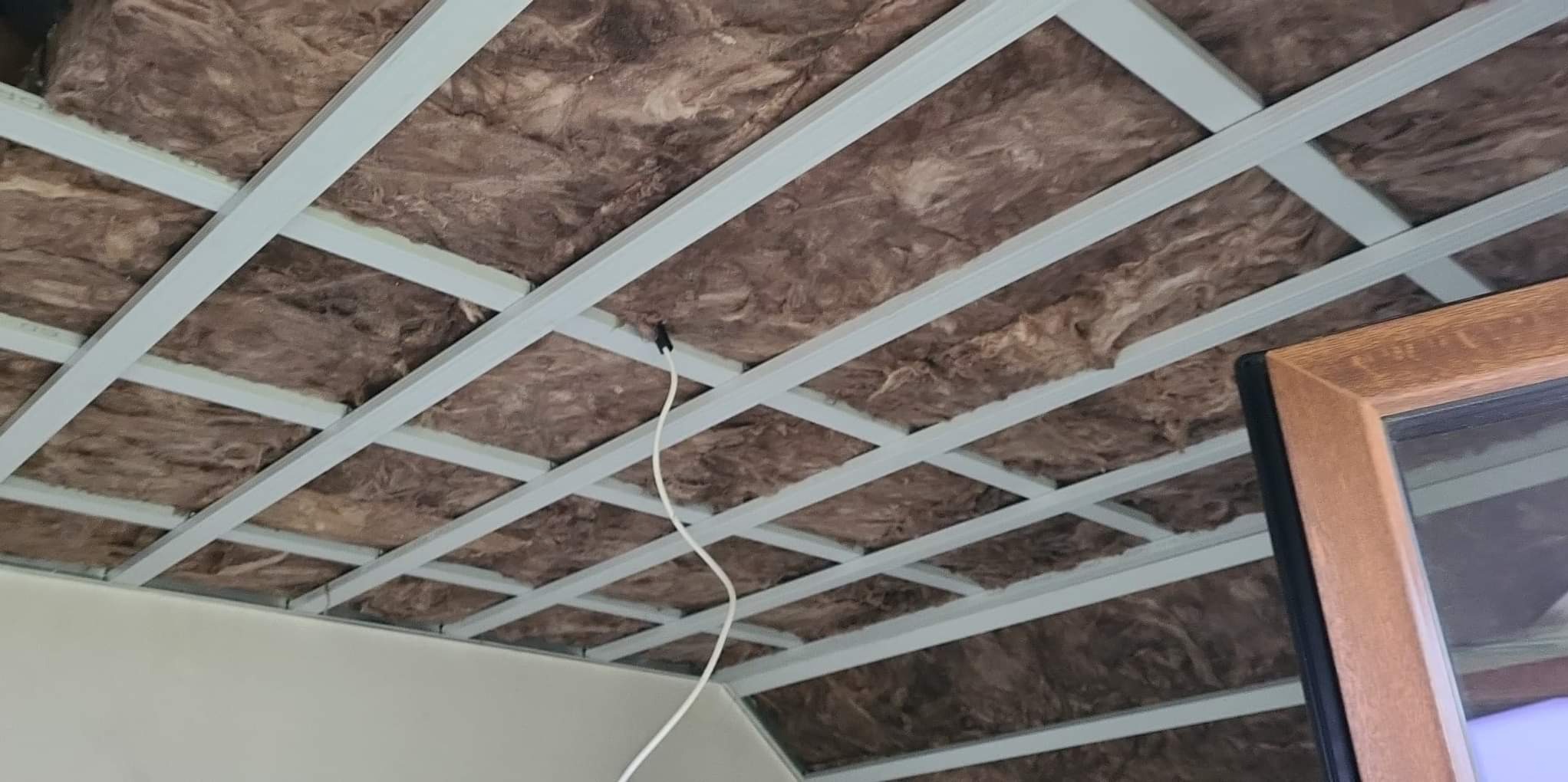 Sufit podwieszany w trakcie montażu, widoczne profile stalowe i izolacja z wełny mineralnej, wystający przewód elektryczny, fragment okna w prawym dolnym rogu.
