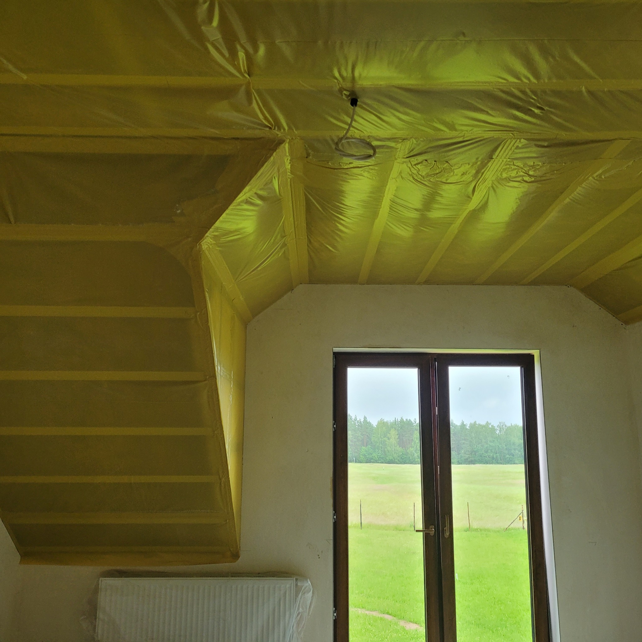 Wnętrze w trakcie remontu: zabezpieczona folią sufitowa konstrukcja drewniana poddasza, widoczny kabel oświetleniowy, okno z widokiem na zielony krajobraz, zakryty folią grzejnik.