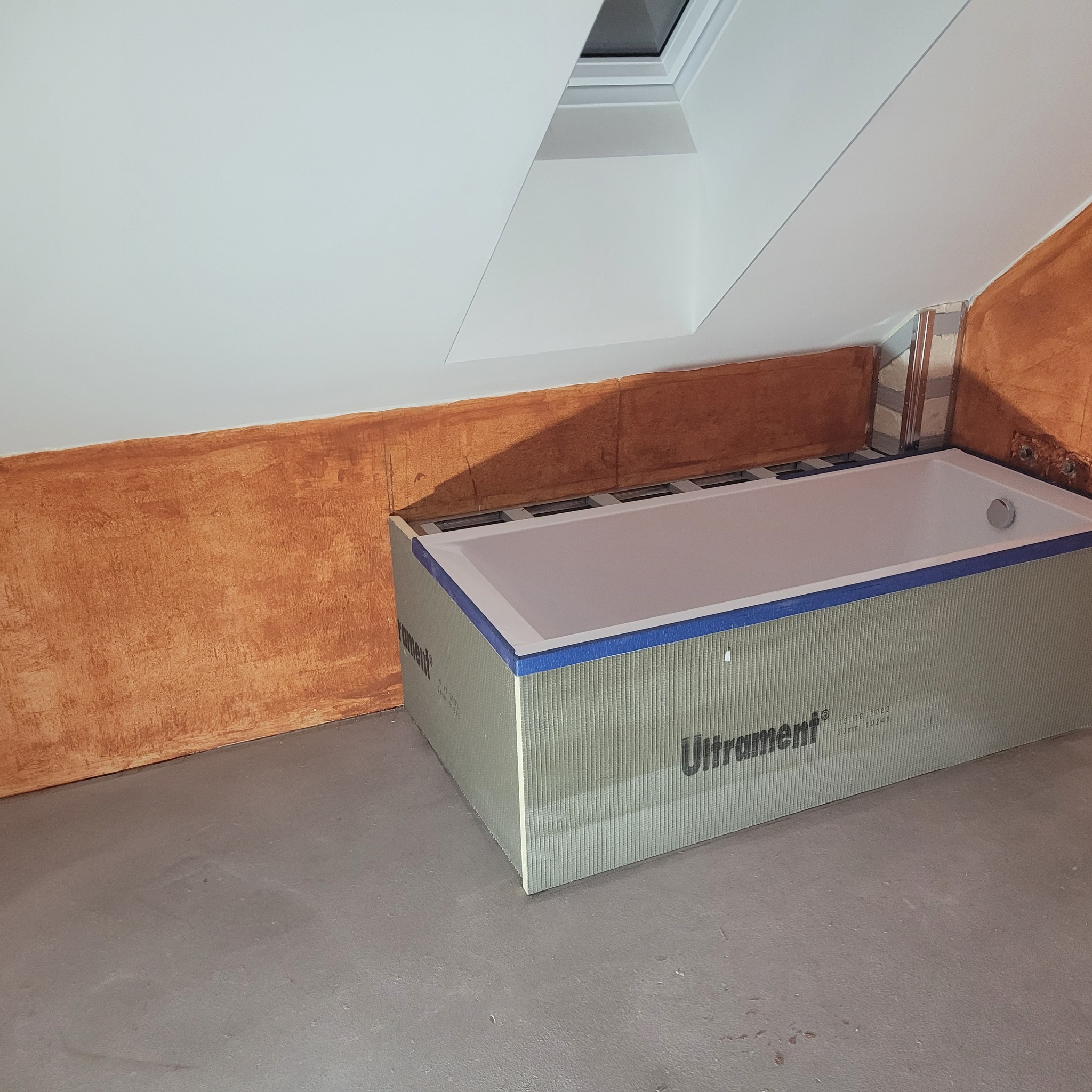 Wanna w obudowie z płyt budowlanych Ultrament, zabezpieczona niebieską taśmą, w trakcie prac wykończeniowych łazienki na poddaszu ze skośnym sufitem i oknem dachowym.