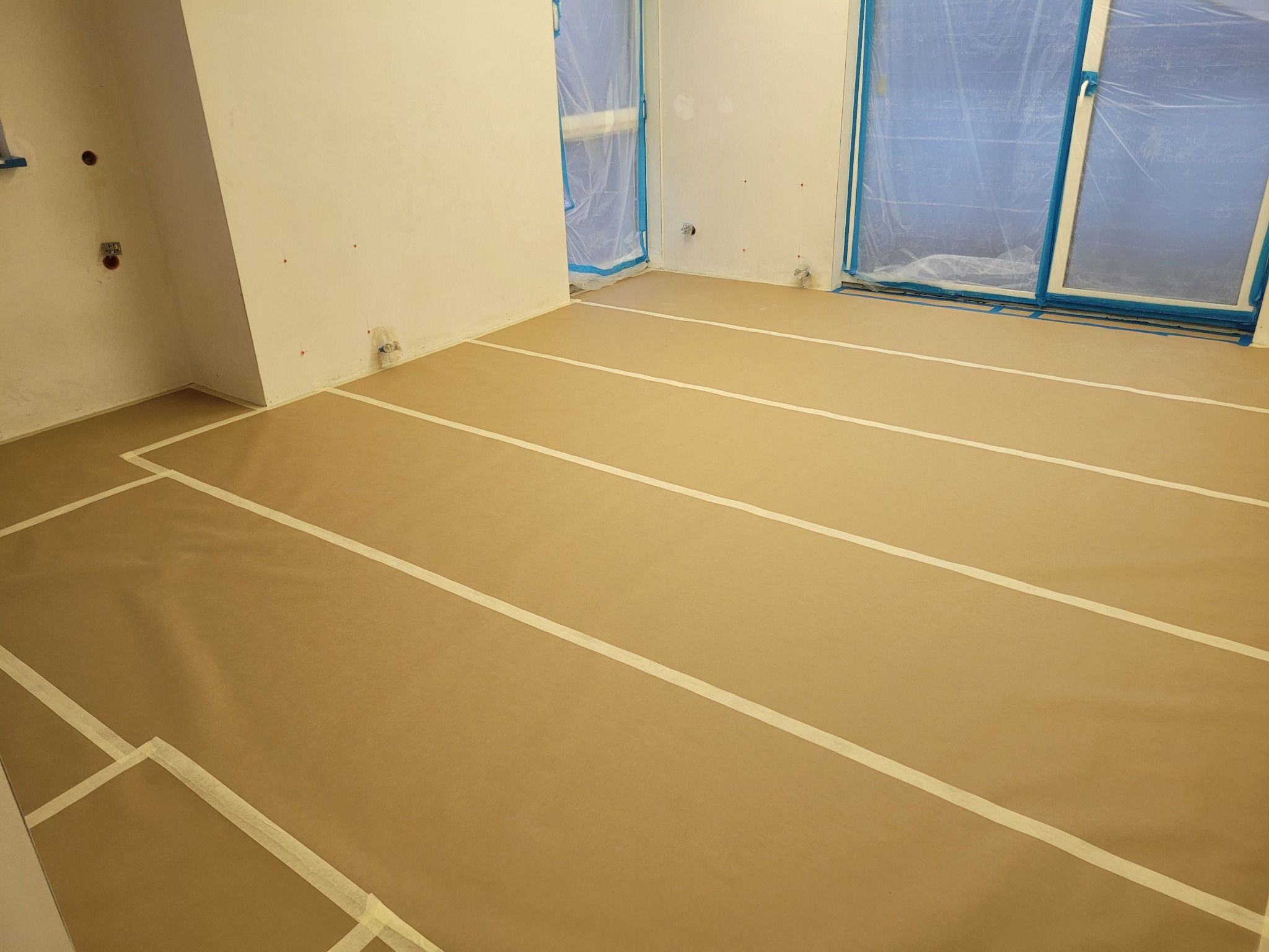 Przygotowanie do malowania: zabezpieczona folią i taśmą podłoga w pustym pokoju z oknami osłoniętymi folią malarską.