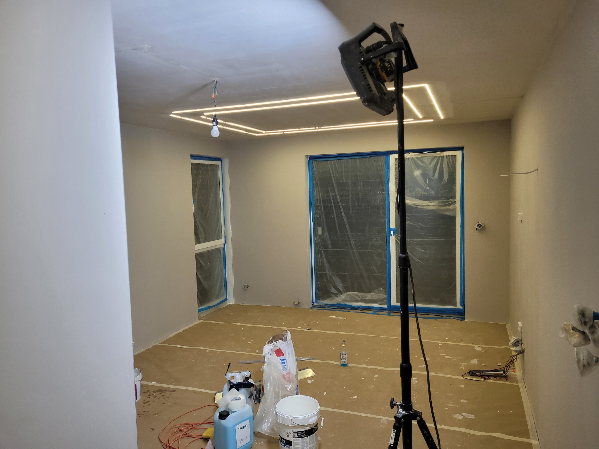 Wnętrze w trakcie prac wykończeniowych: zabezpieczone okna folią, podłoga wyłożona kartonem, widoczny sprzęt oświetleniowy na statywie, puszki z farbą i akcesoria malarskie, oraz zamontowane...
