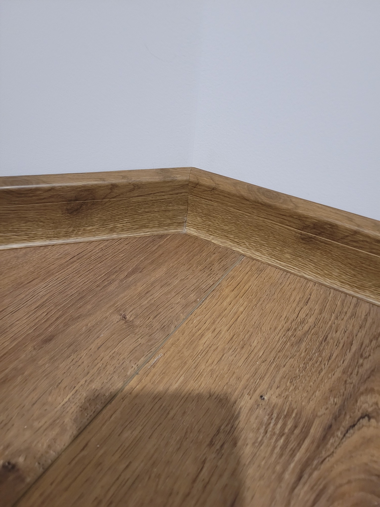 Fragment podłogi z paneli w odcieniach brązu, z widoczną listwą przypodłogową w dopasowanym kolorze, przylegającą do białej ściany w rogu pomieszczenia.