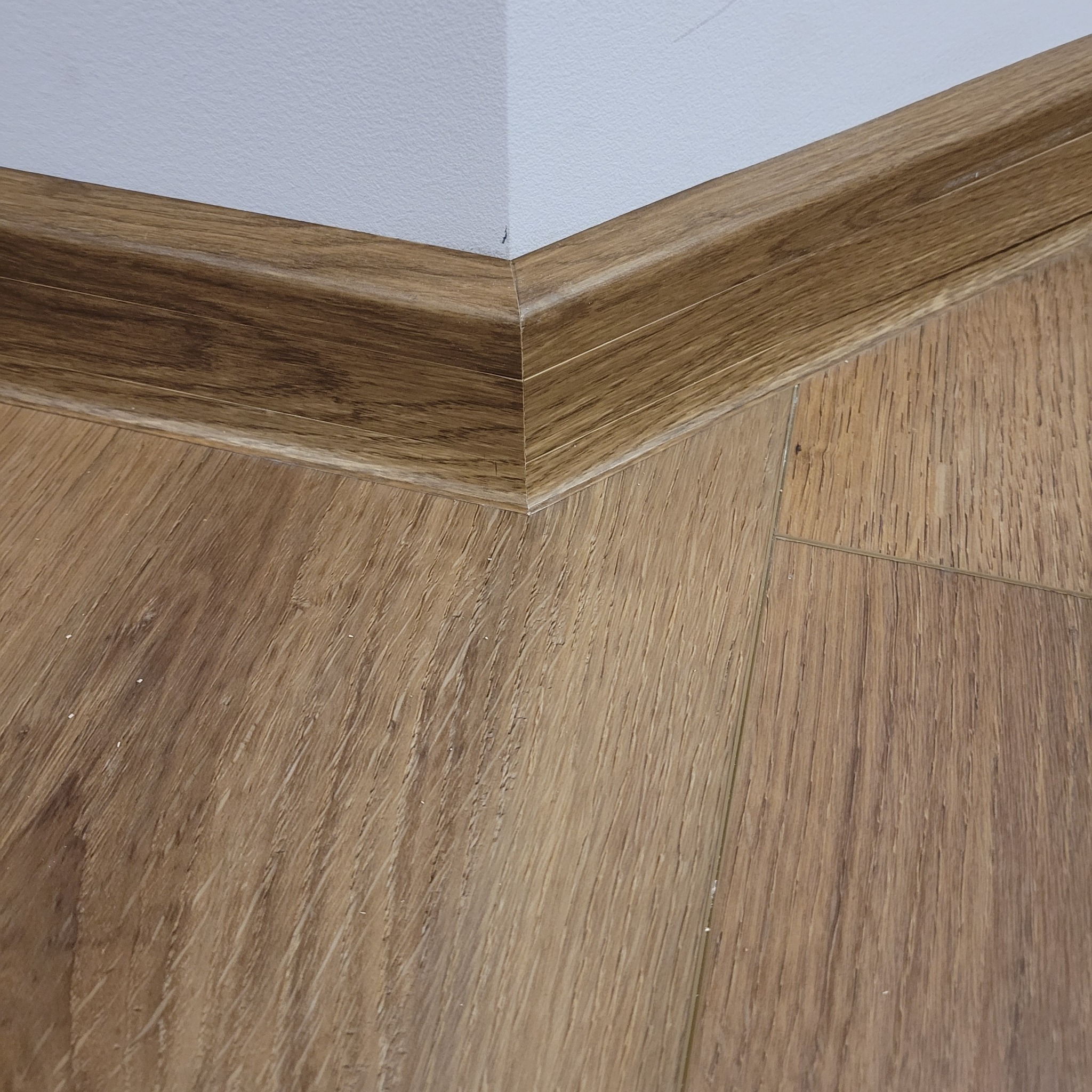 Drewnopodobna podłoga laminowana z dopasowaną listwą przypodłogową w rogu pomieszczenia, widoczne łączenia paneli.