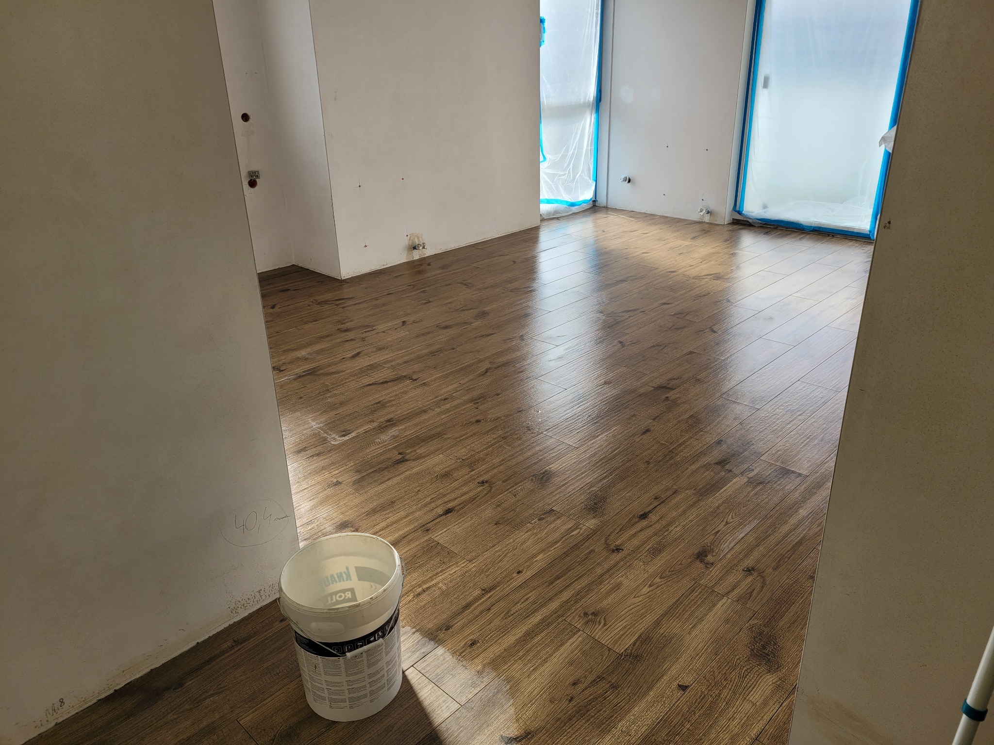 Ukończona podłoga z paneli drewnopodobnych w nowym mieszkaniu, zabezpieczone okna folią malarską, widoczne ślady po pomiarach na ścianie i wiadro z farbą Knauf.