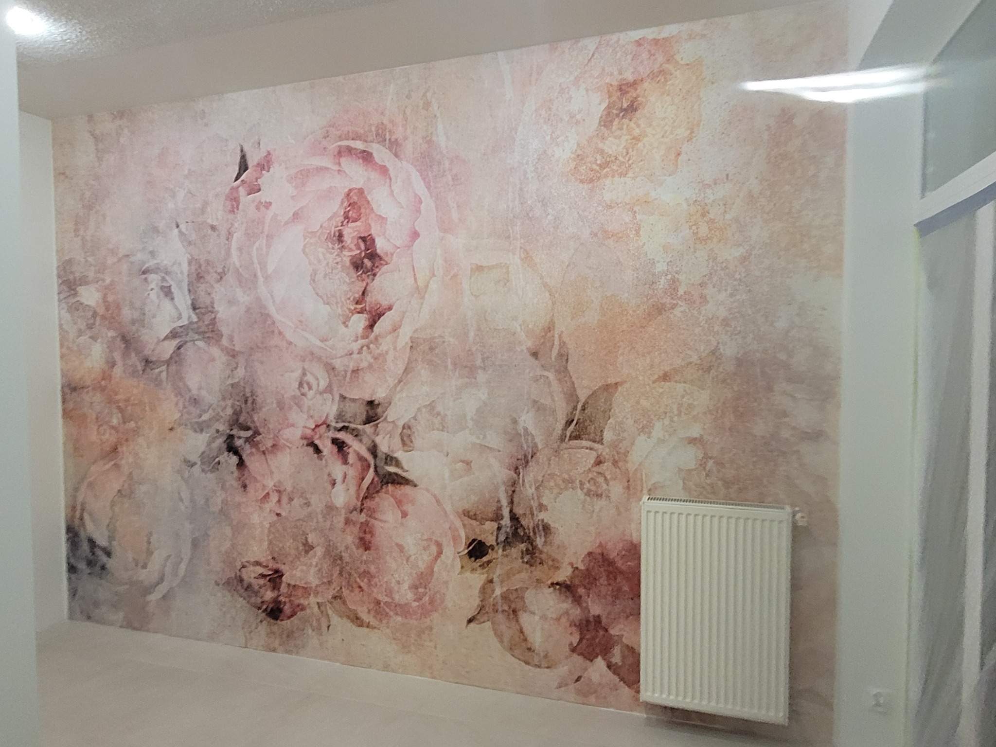 Ściana z fototapetą w kwiaty w odcieniach różu i beżu, grzejnik na ścianie, widok z perspektywy pokoju, delikatne pastelowe barwy.