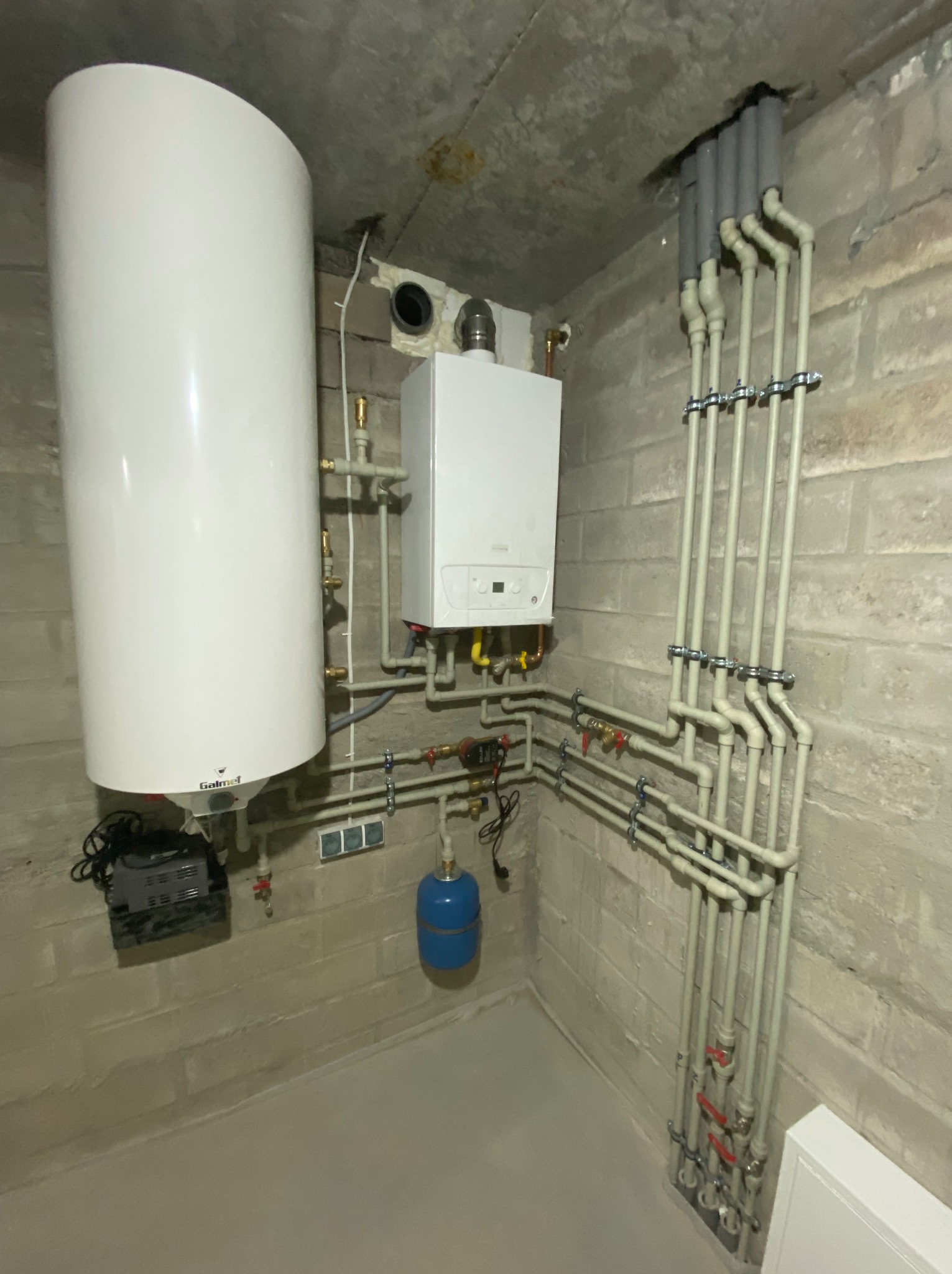 Biały piec gazowy i zasobnik ciepłej wody z kompletnym systemem rur na ścianie z betonu komórkowego, widoczne elementy instalacji i podłączenia.
