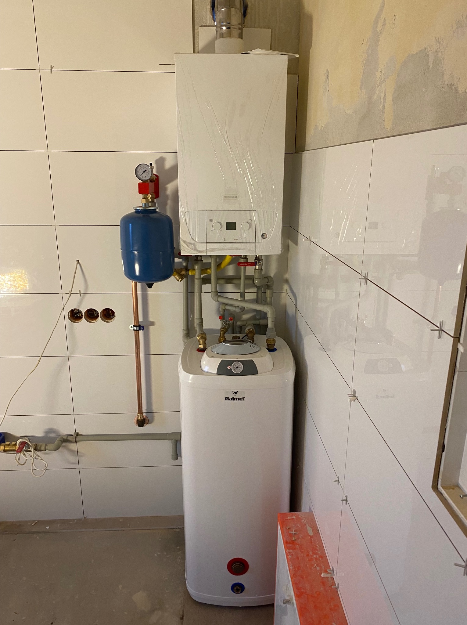 Biały kondensacyjny piec gazowy marki De Dietrich i zbiornik Galmet, z niebieskim naczyniem przeponowym. Białe płytki na ścianach, widoczne ślady po instalacji.