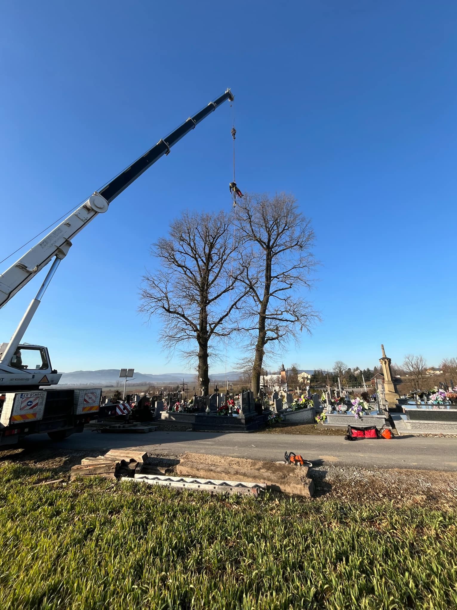 Wycinka drzew na cmentarzu: pracownik na linie, podnoszony dźwigiem, obok leżą pocięte fragmenty pnia, a w tle nagrobki i błękitne niebo.