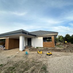 Liubomyr Hrechaniuk - Dom jednorodzinny z szarą dachówką i drewnianą okładziną elewacyjną w trakcie prac wykończeniowych, na pierwszym planie zabawki dziecięce na piasku, w tle dwóch pracowników przy wykopie.