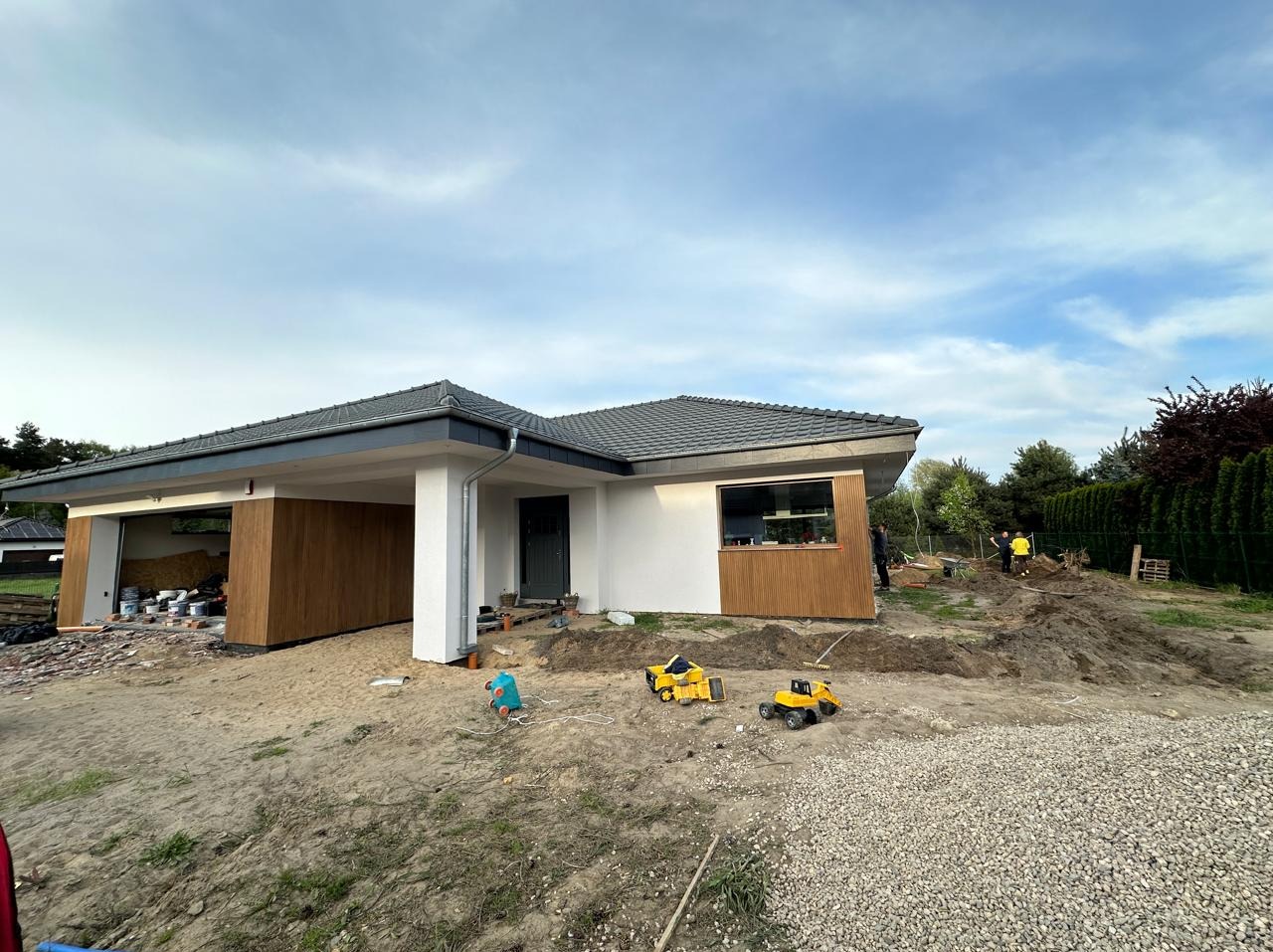 Dom jednorodzinny z szarą dachówką i drewnianą okładziną elewacyjną w trakcie prac wykończeniowych, na pierwszym planie zabawki dziecięce na piasku, w tle dwóch pracowników przy wykopie.