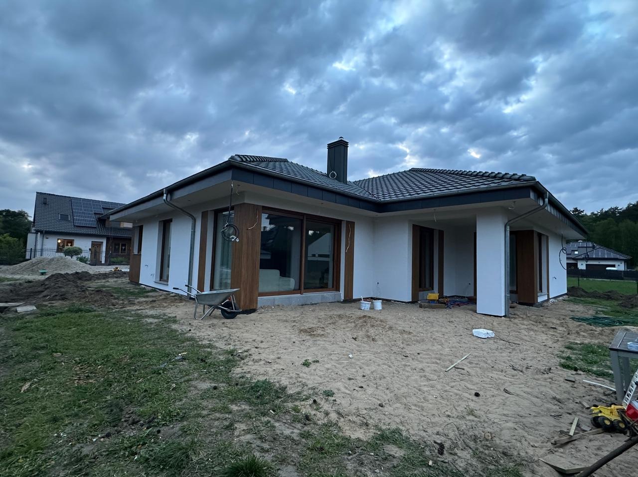 Nowoczesny dom jednorodzinny z białymi ścianami i ciemną stolarką okienną, w trakcie prac wykończeniowych elewacji, widoczny wózek budowlany i narzędzia na piaszczystym terenie.