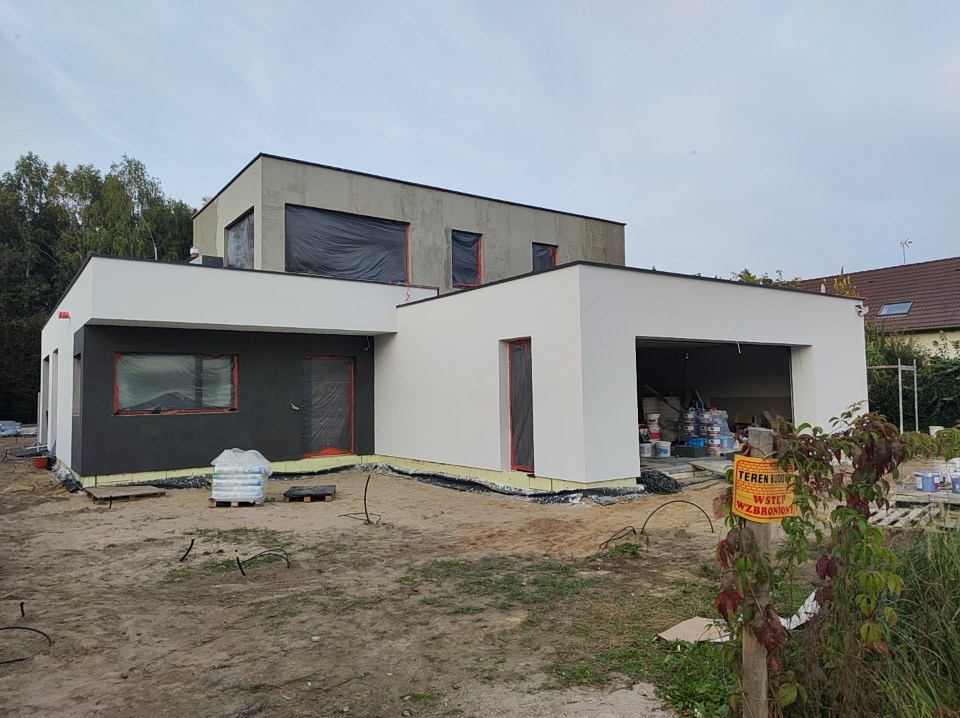 Nowoczesny dom w trakcie prac elewacyjnych: część ściany pomalowana na biało, część na grafitowo, okna zabezpieczone folią, widoczne worki z materiałami budowlanymi i farby, tablica informacyjna...