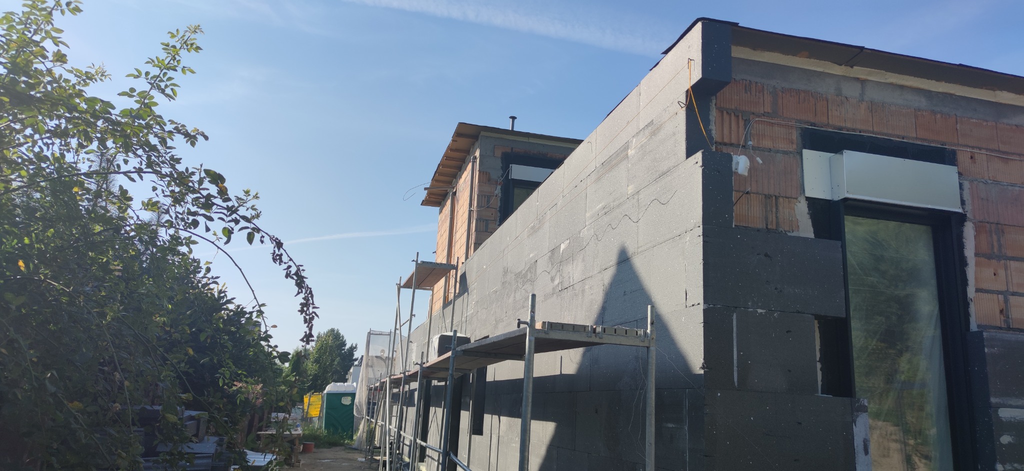 Dom w trakcie ocieplania styropianem grafitowym, widoczne rusztowania i fragmenty niezabezpieczonej cegły, w tle błękitne niebo i zieleń drzew.