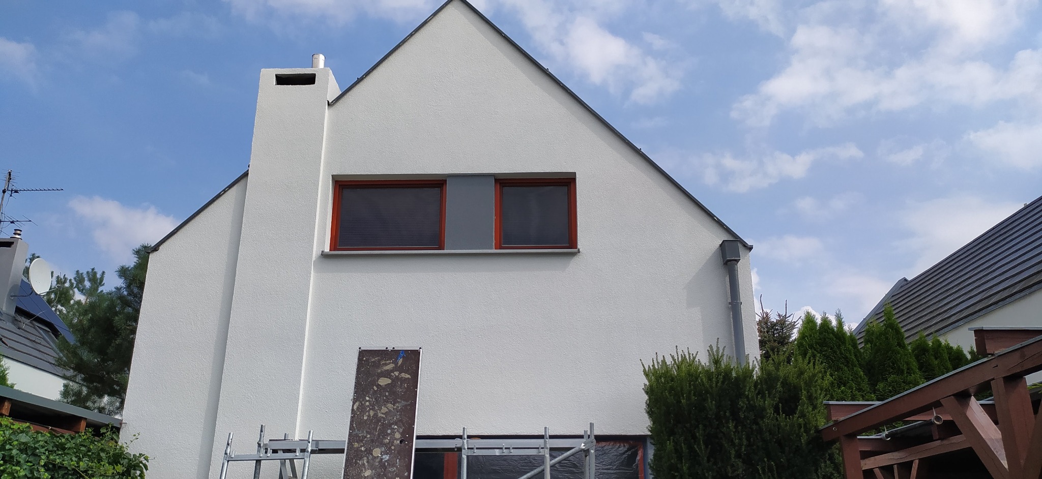 Biała elewacja domu jednorodzinnego z dwoma oknami w brązowych ramach, fragment rusztowania i osłonięte folią okno w dolnej części, widok na błękitne niebo z chmurami.