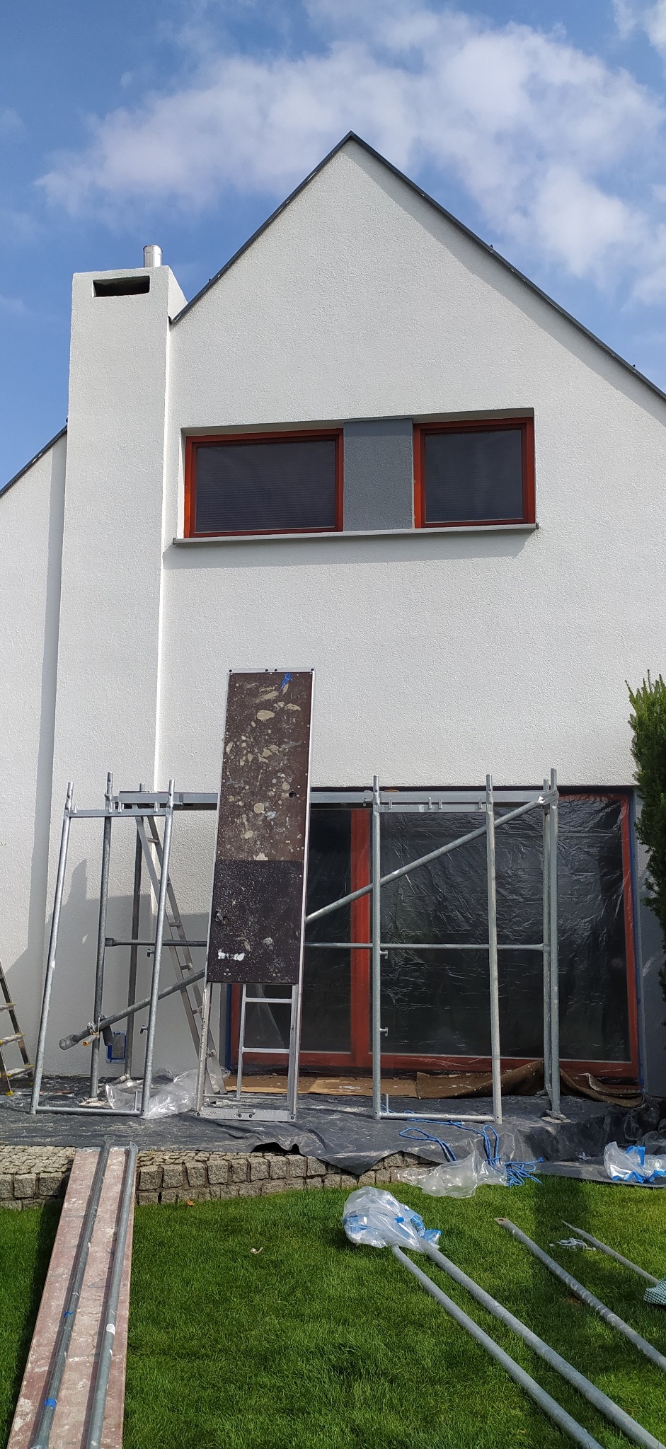 Dom jednorodzinny z rusztowaniem przed oknem zabezpieczonym folią, przygotowany do prac na elewacji. Widoczna deska z resztkami zaprawy oraz elementy rusztowania leżące na trawniku.