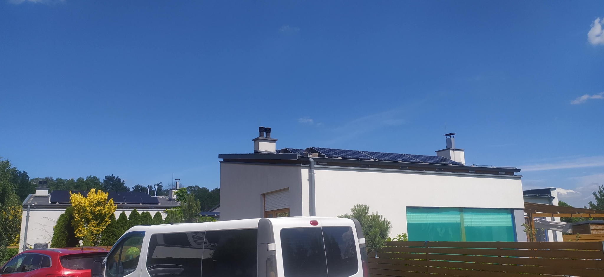 Biały dom z panelami słonecznymi na dachu, widoczny zza białego busa i częściowo zasłonięty przez drewniane ogrodzenie, na tle błękitnego nieba.